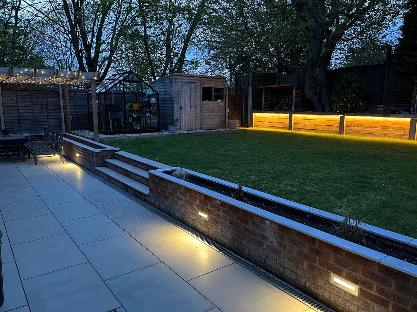Landscaped garden at dusk with a porcelain patio, raised brick beds and recessed lighting by DNR Build