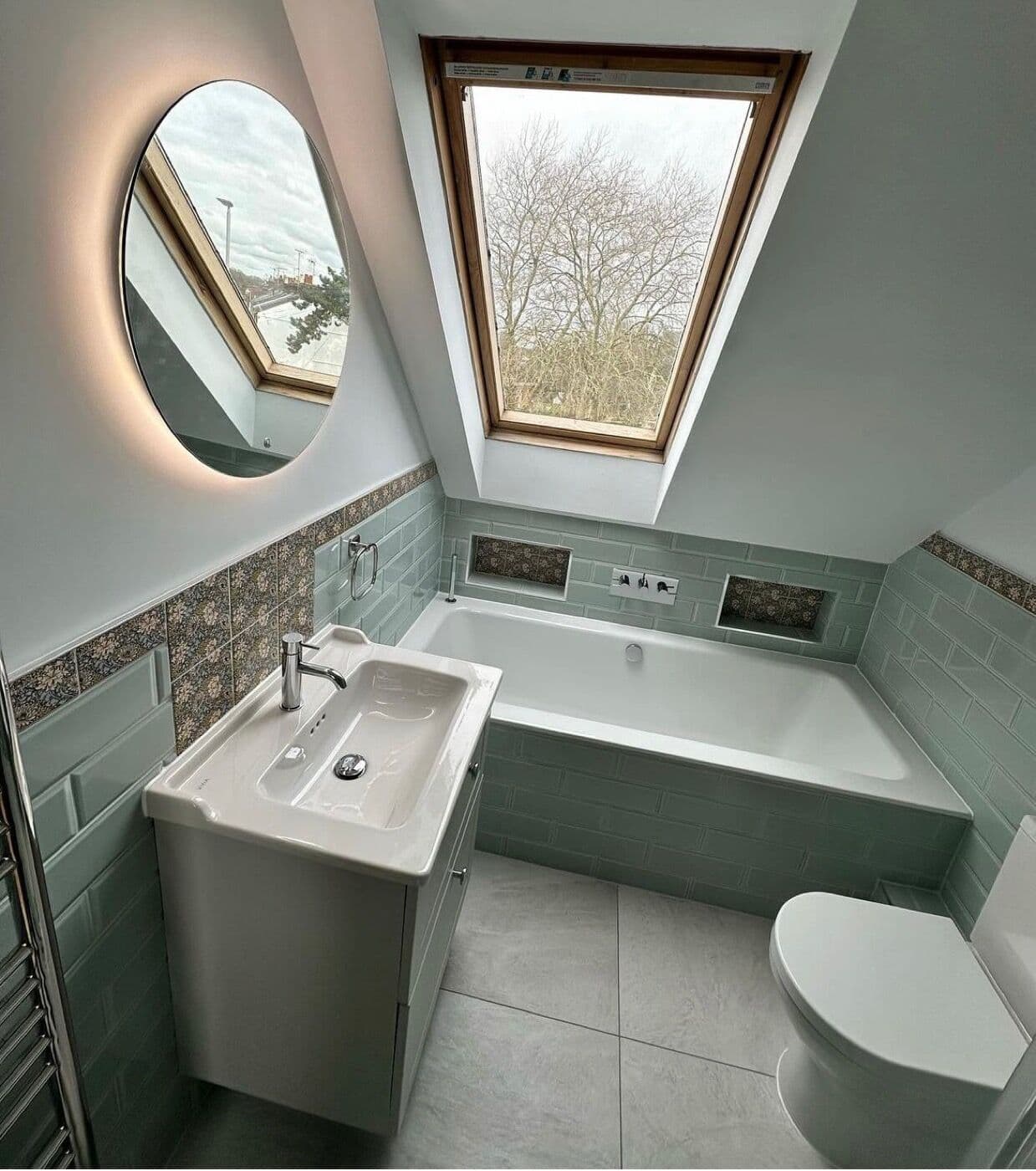 Renovated loft bathroom with sage tiling, a bath under a roof window and backlit mirror by DNR Build
