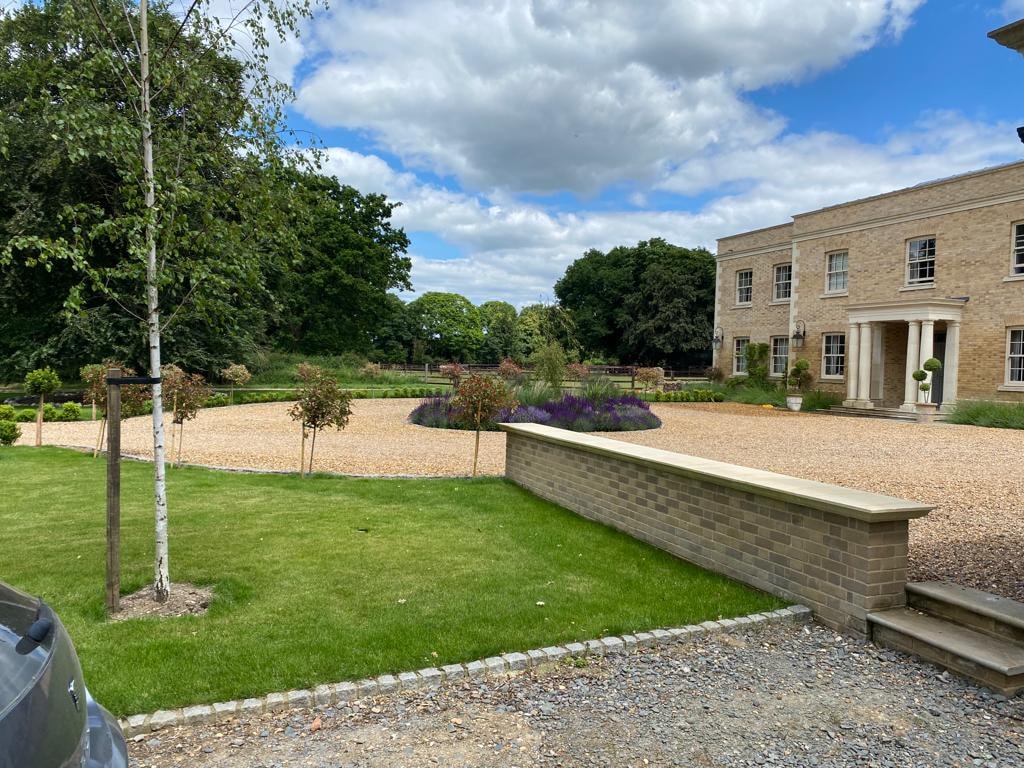 Landscaped grounds with a brick retaining wall, lawns and planted borders beside the country house by DNR Build