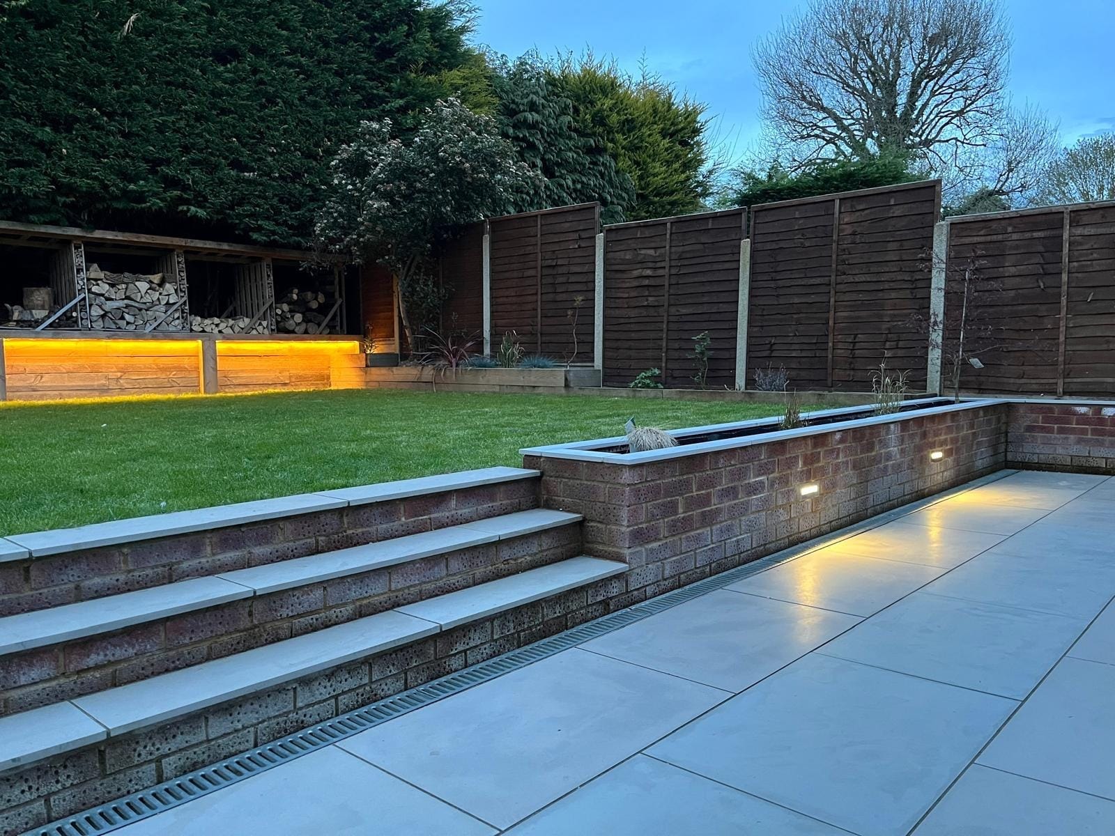 The lit patio at dusk, with porcelain steps and a brick planter wall leading up to the lawn and the glowing sleeper wall beyond by DNR Build