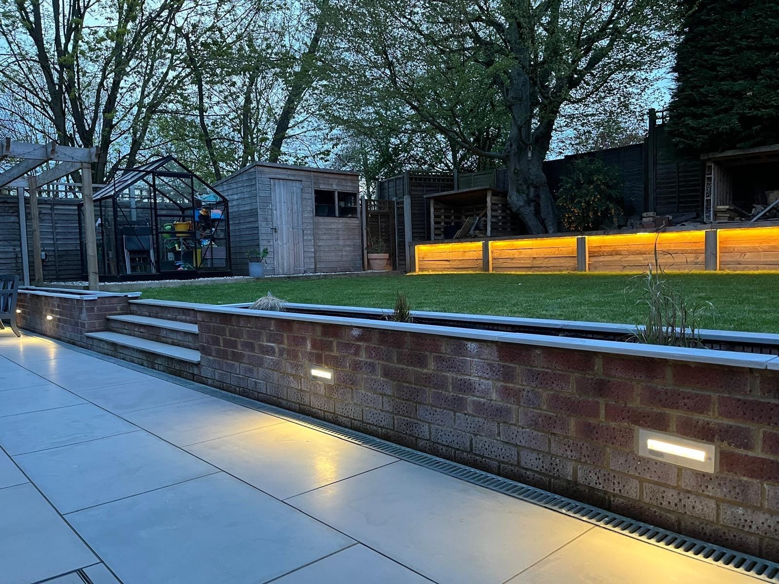 Brick planter wall with built in lights along the porcelain patio, looking towards the greenhouse, shed and lit sleeper wall by DNR Build
