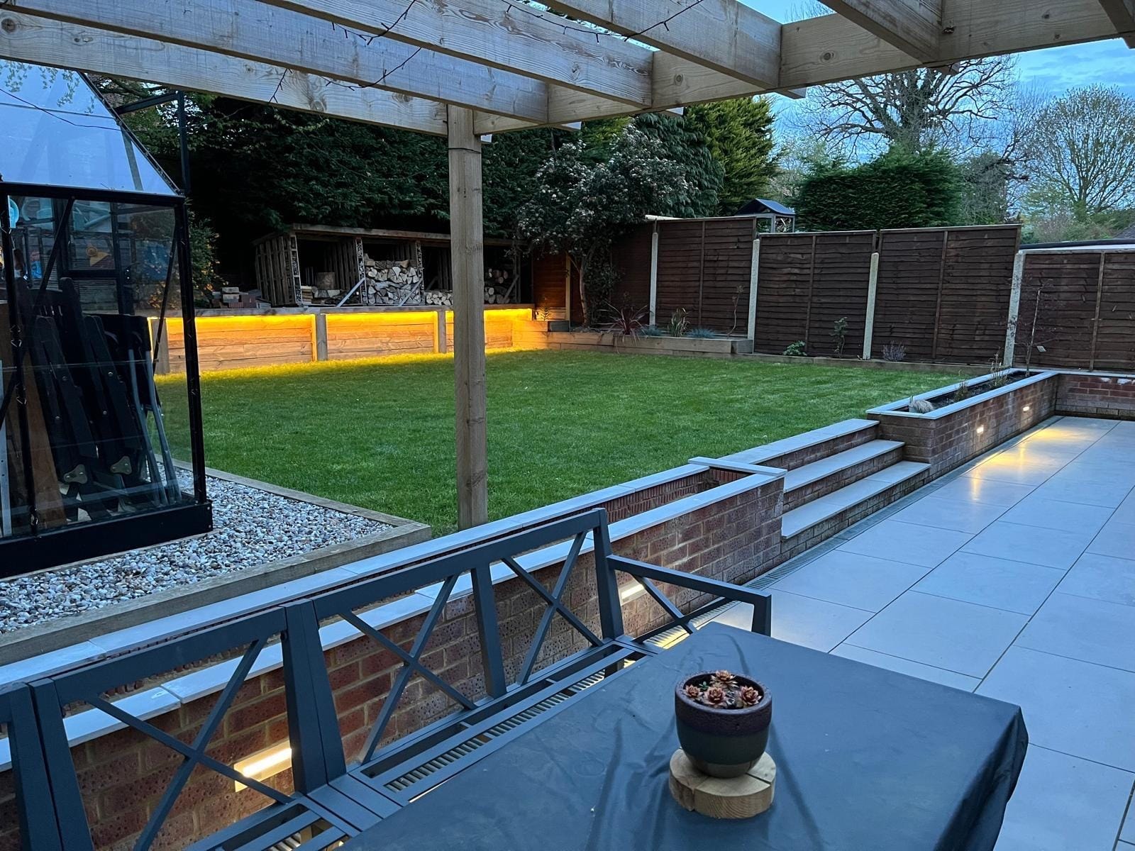 View from under the pergola across the lawn to the lit sleeper wall and log stores, with the dining set in the foreground by DNR Build