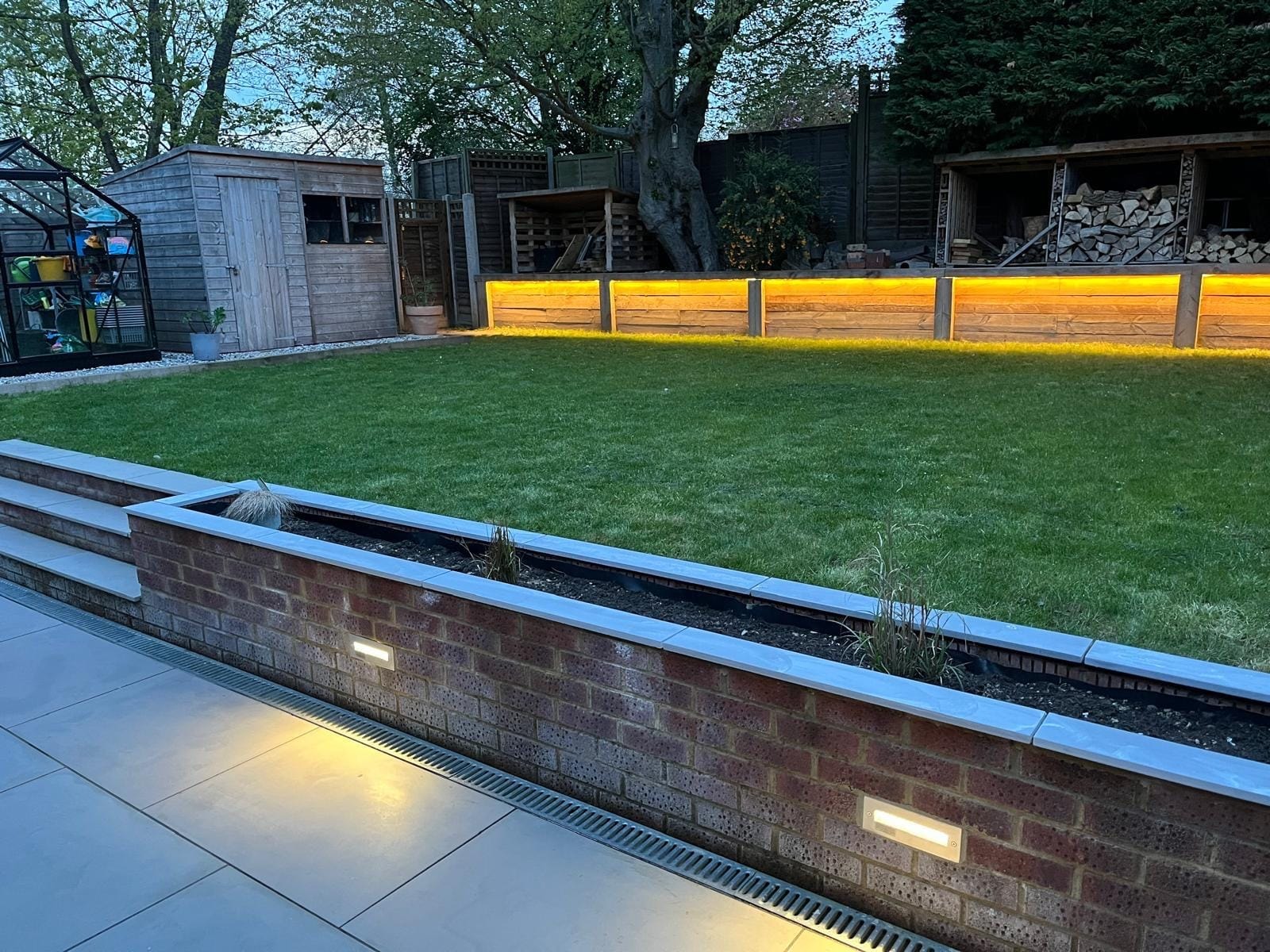 Lit brick planter wall alongside the patio drainage channel, with the lawn and glowing sleeper wall behind by DNR Build