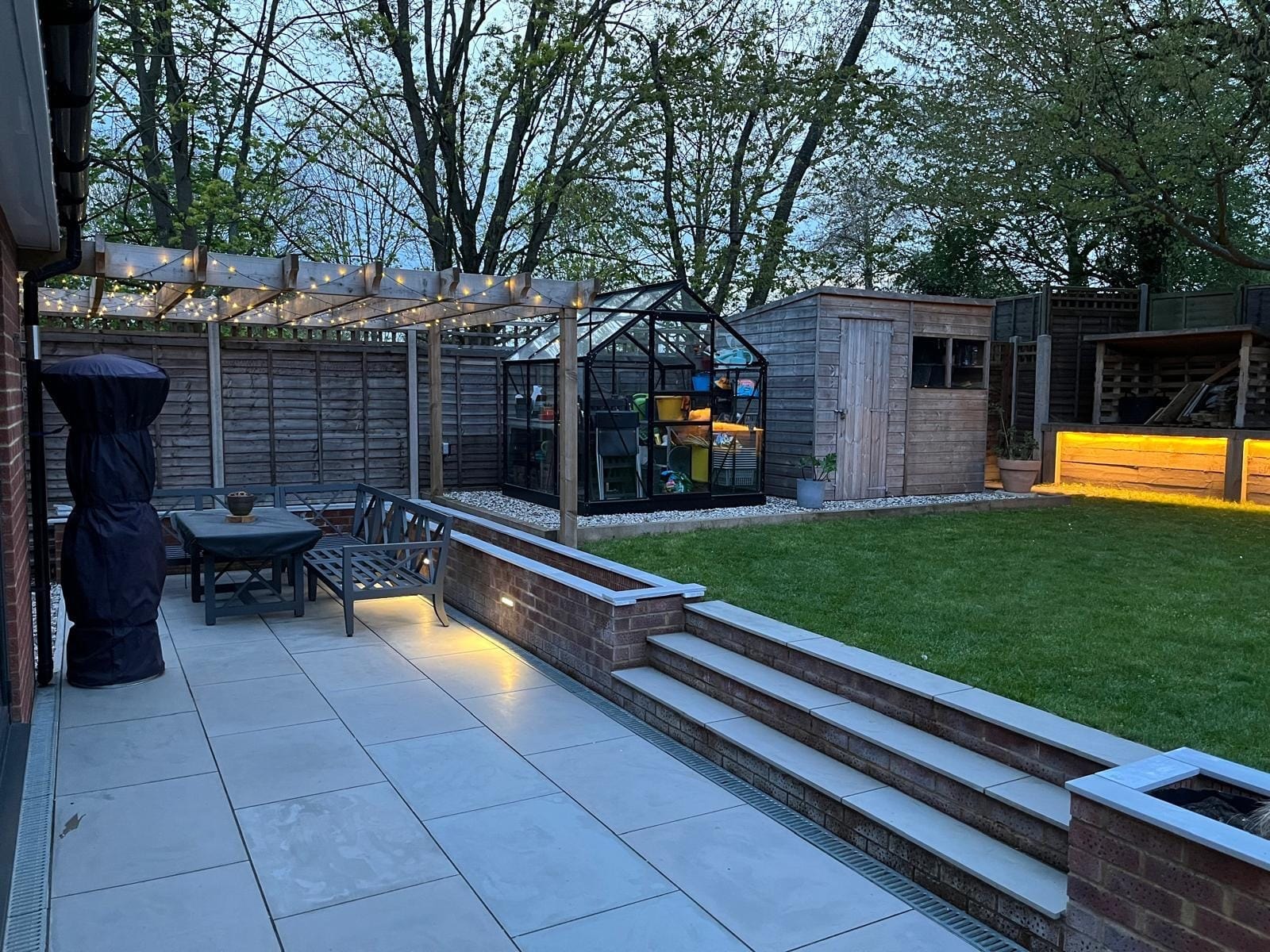 The patio running from the house past the pergola seating area, with steps up to the lawn and the lights on by DNR Build