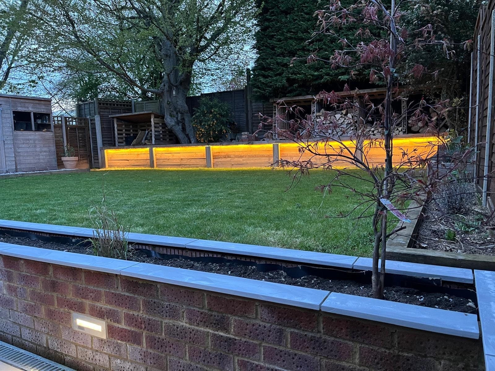 Planted brick wall in the foreground with the lawn and the warm glow of the sleeper retaining wall beyond by DNR Build