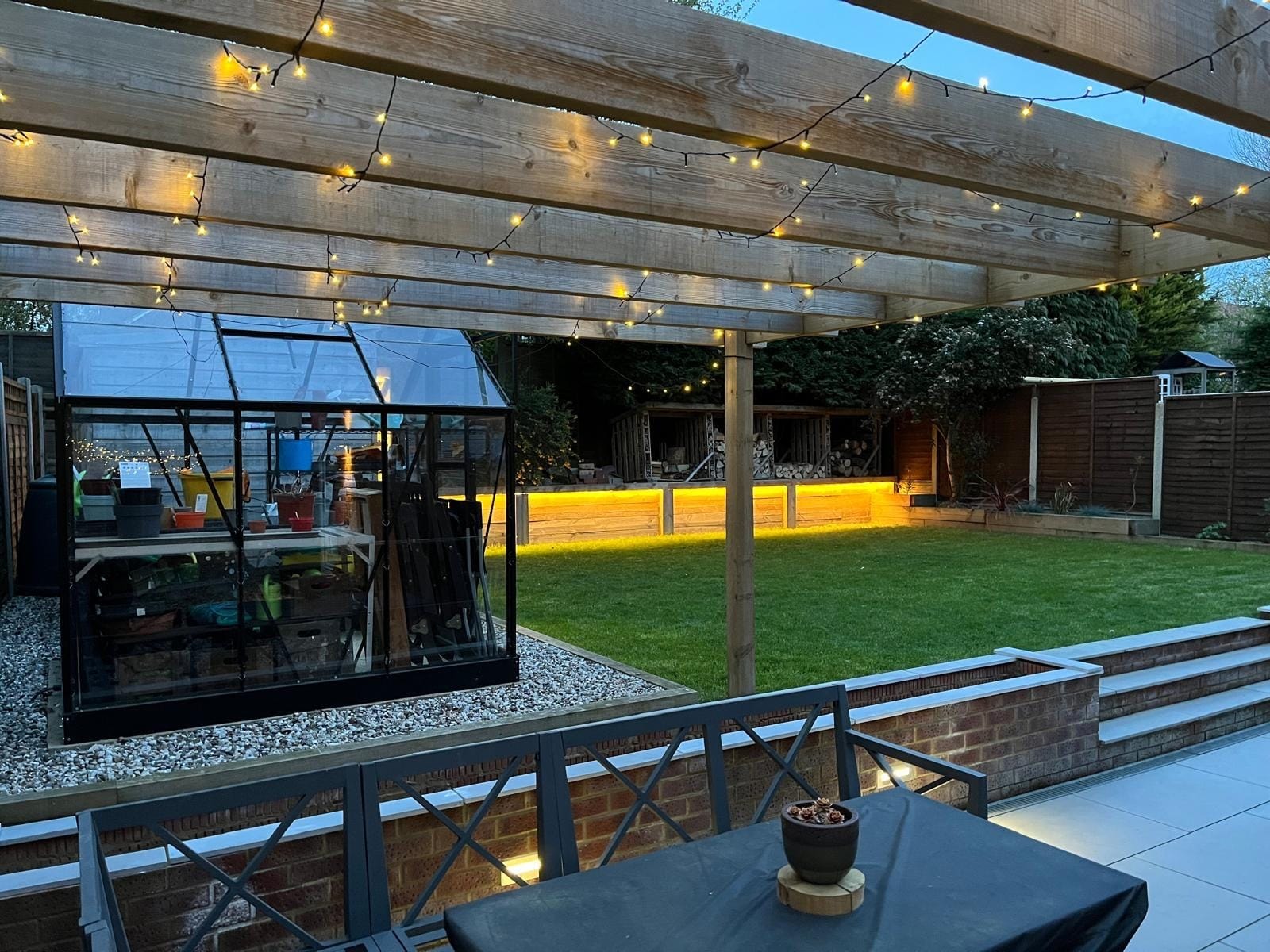 Festoon lights strung under the timber pergola, looking out to the greenhouse and the lit sleeper wall by DNR Build