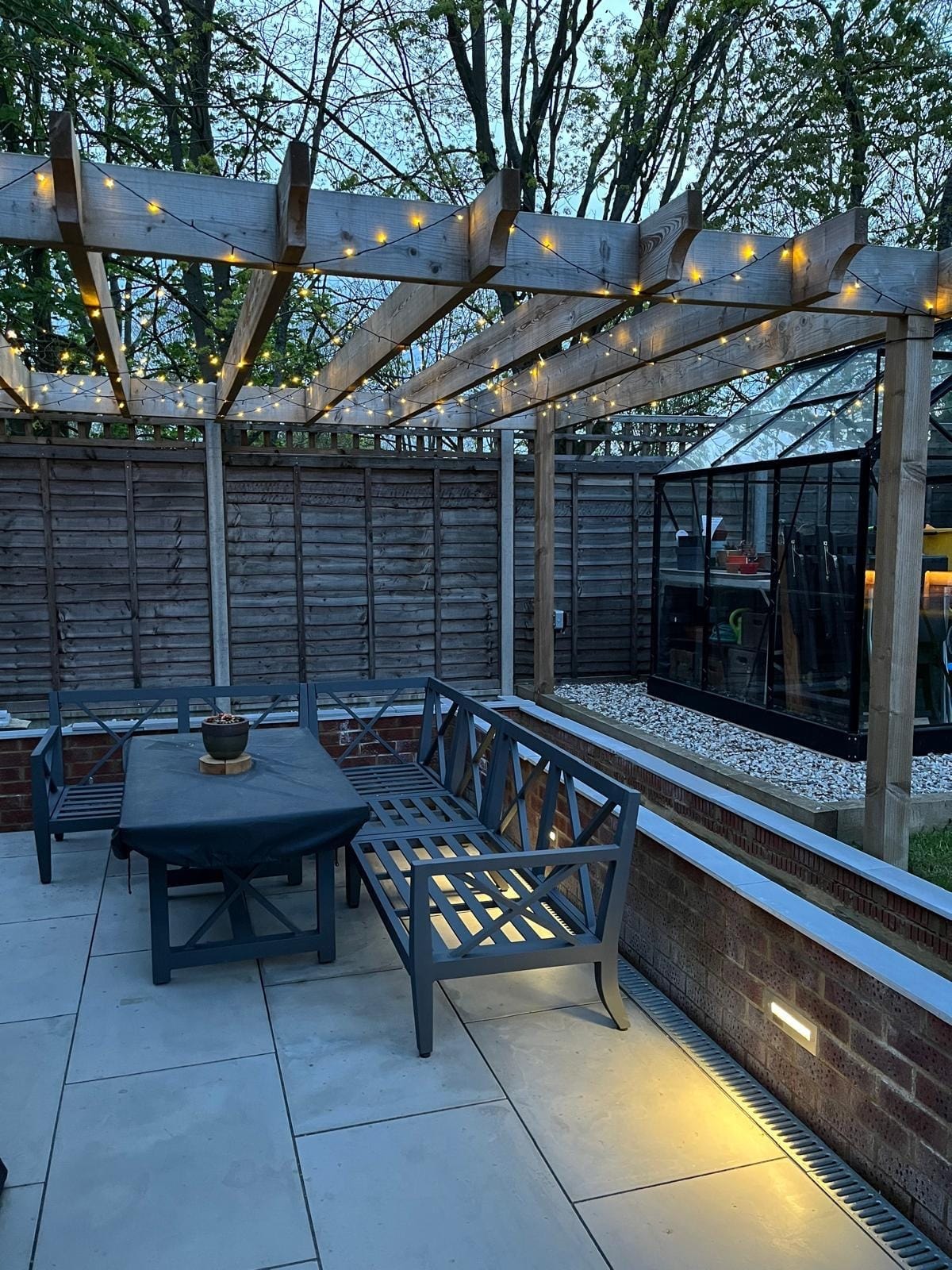 The seating area under the lit pergola, with the brick planter wall and greenhouse alongside by DNR Build