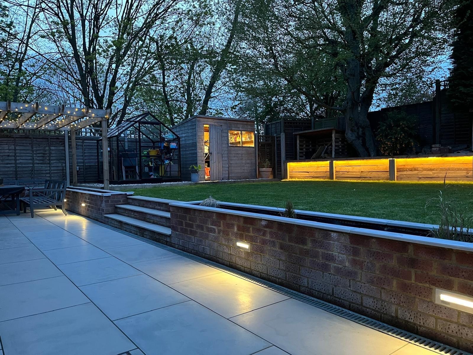 Wide view along the lit patio and planter wall to the pergola, greenhouse, shed and sleeper wall by DNR Build