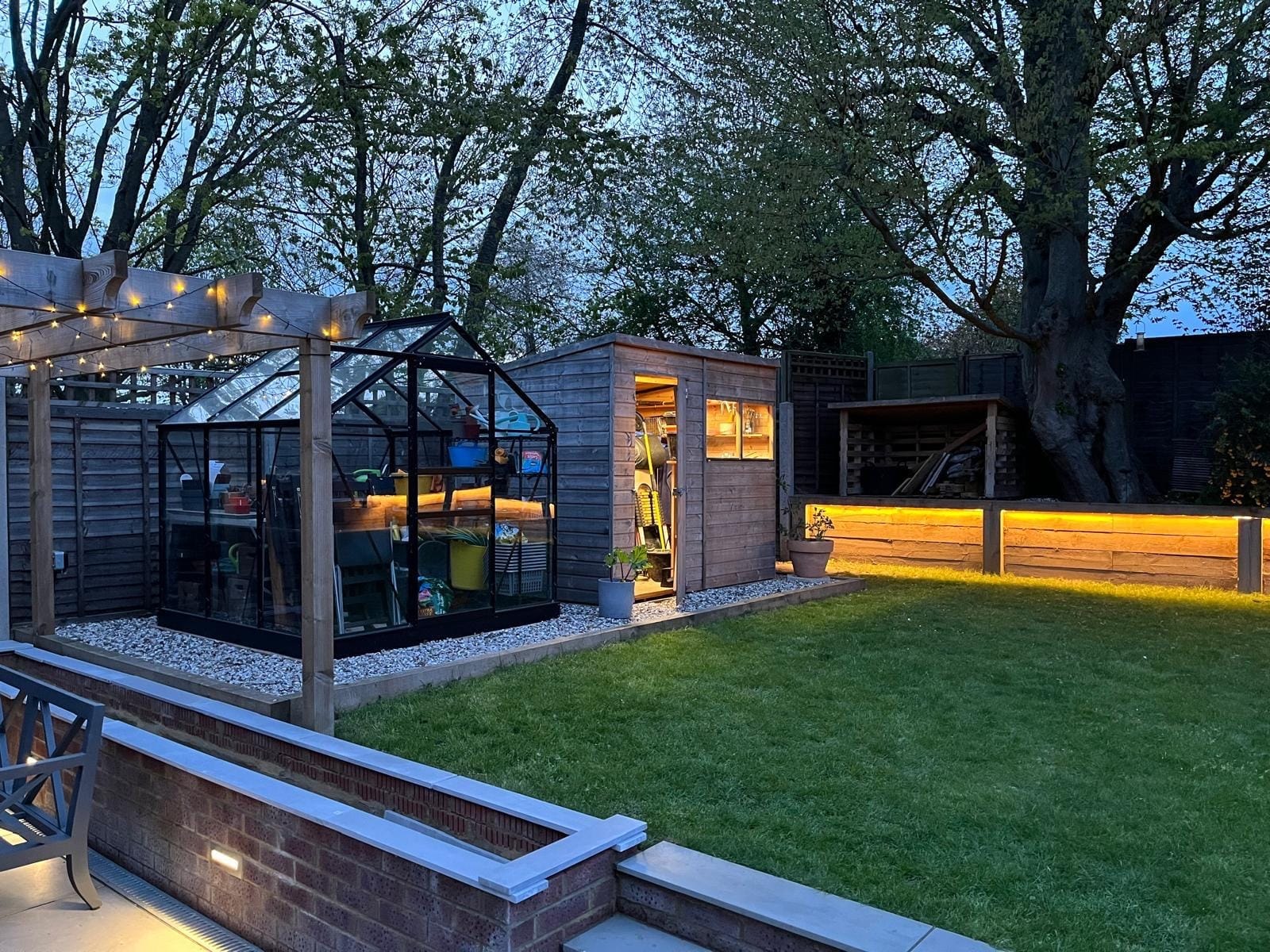 The greenhouse and shed on their gravel base beside the lawn, with the sleeper wall lit at the far end by DNR Build