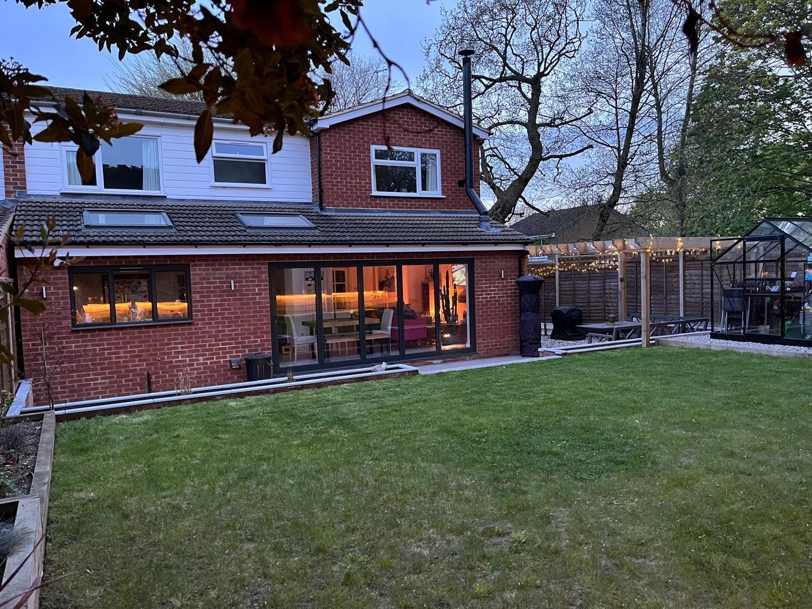 The rear of the house with its bi-fold doors and the finished lawn, pergola and greenhouse at dusk by DNR Build