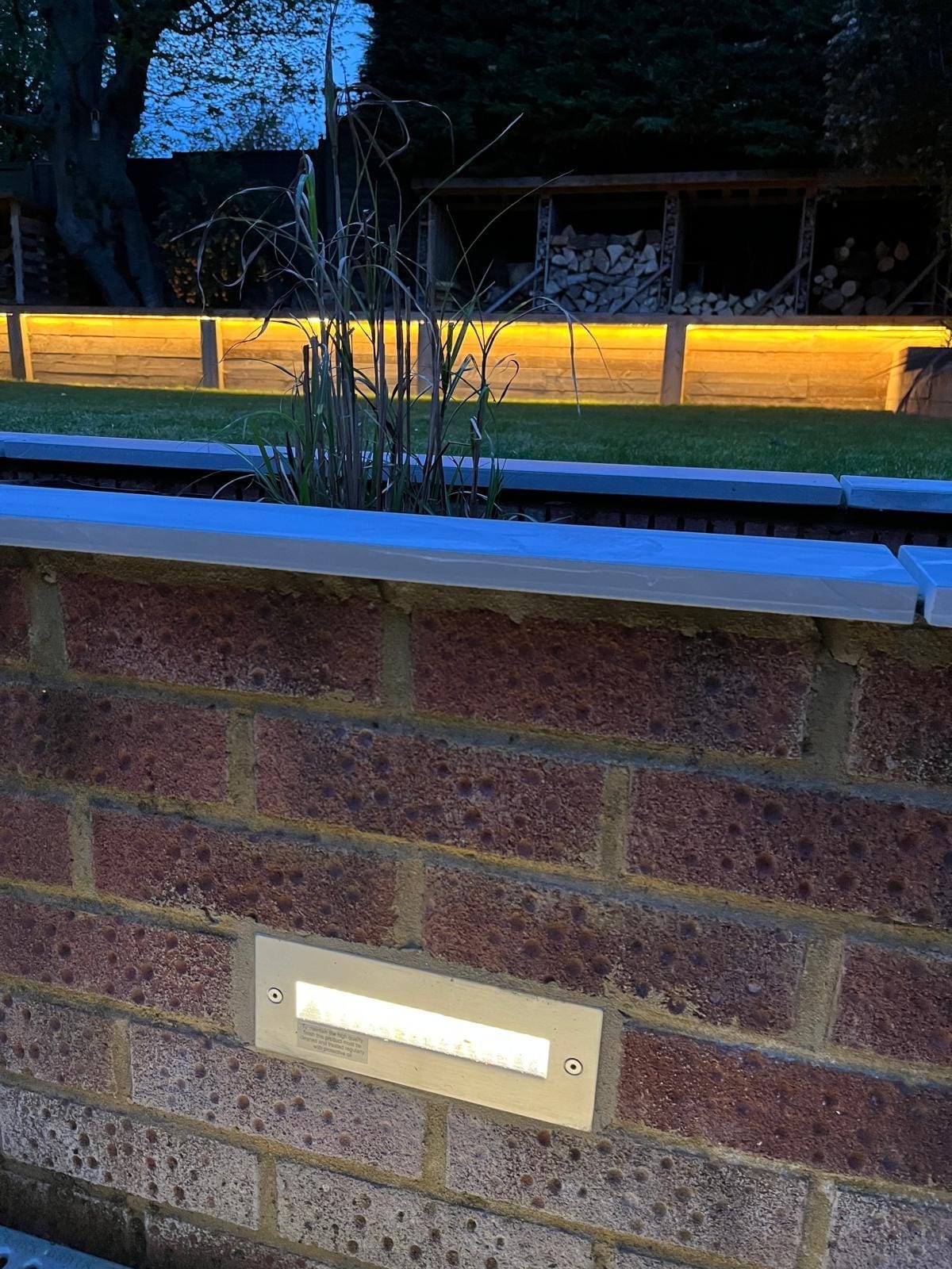 Close up of a recessed light in the brick planter wall, with the lit sleeper wall across the lawn by DNR Build