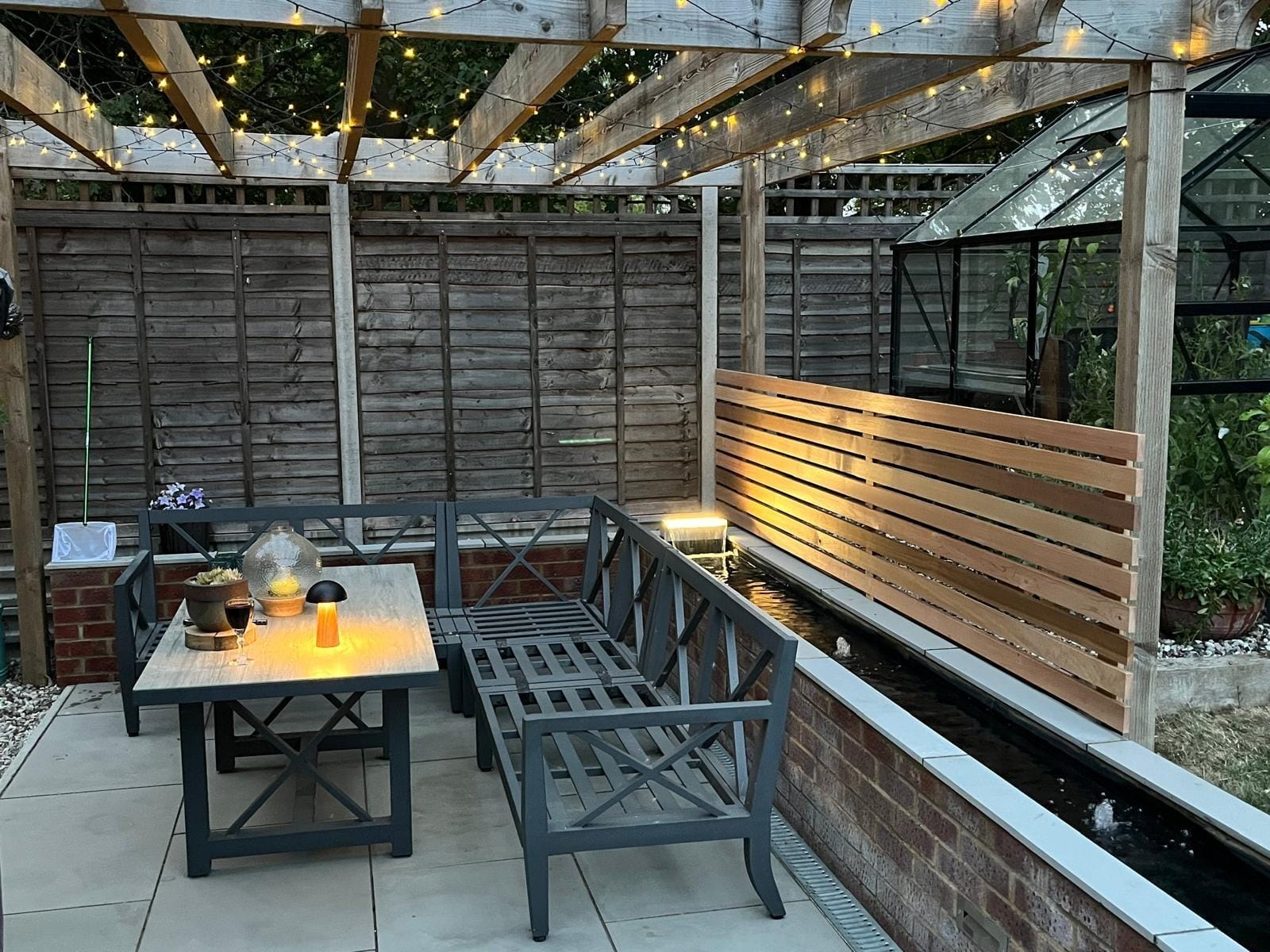 The sheltered seating corner under the pergola, with a slatted cedar screen and a lit water feature along the wall by DNR Build