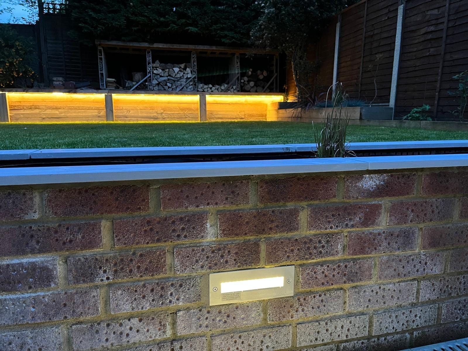 A recessed step light in the brick wall, with the glowing sleeper wall and log stores beyond the lawn by DNR Build