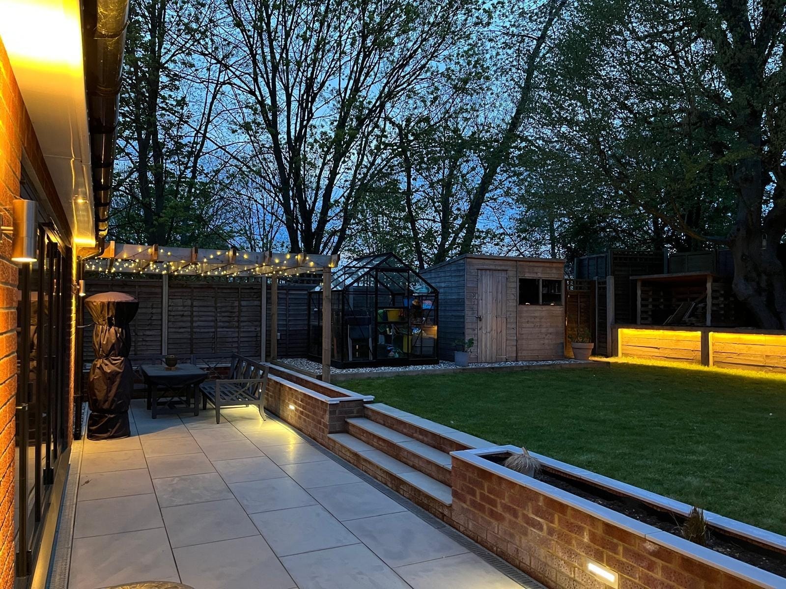 Looking along the patio from the house, with wall lights, the pergola seating area and the lit planter wall by DNR Build