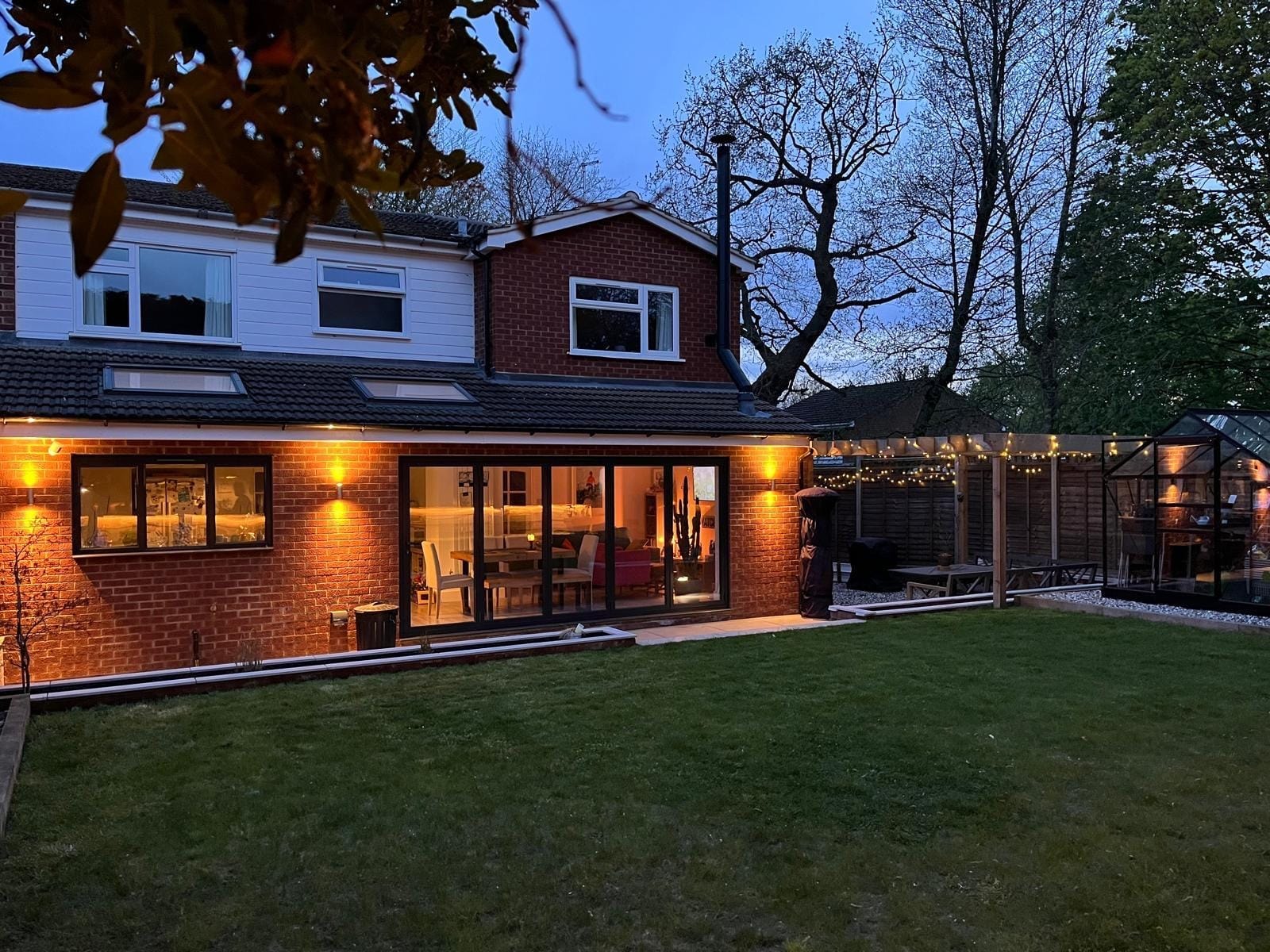 The finished extension and garden at dusk, with warm wall lights and the festoon lit pergola by DNR Build