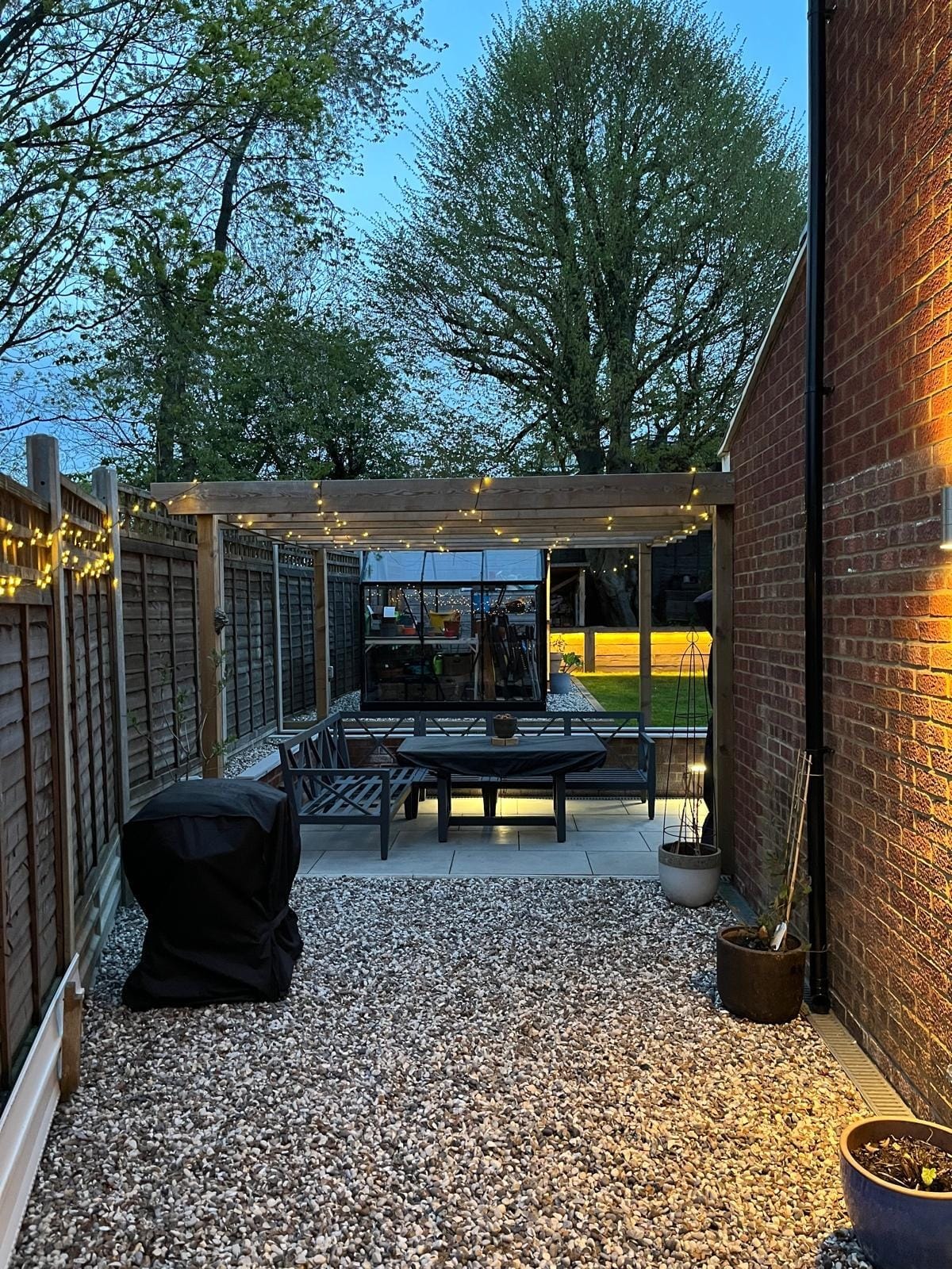 The gravel side garden leading to the pergola seating area, with festoon lights and the greenhouse beyond by DNR Build