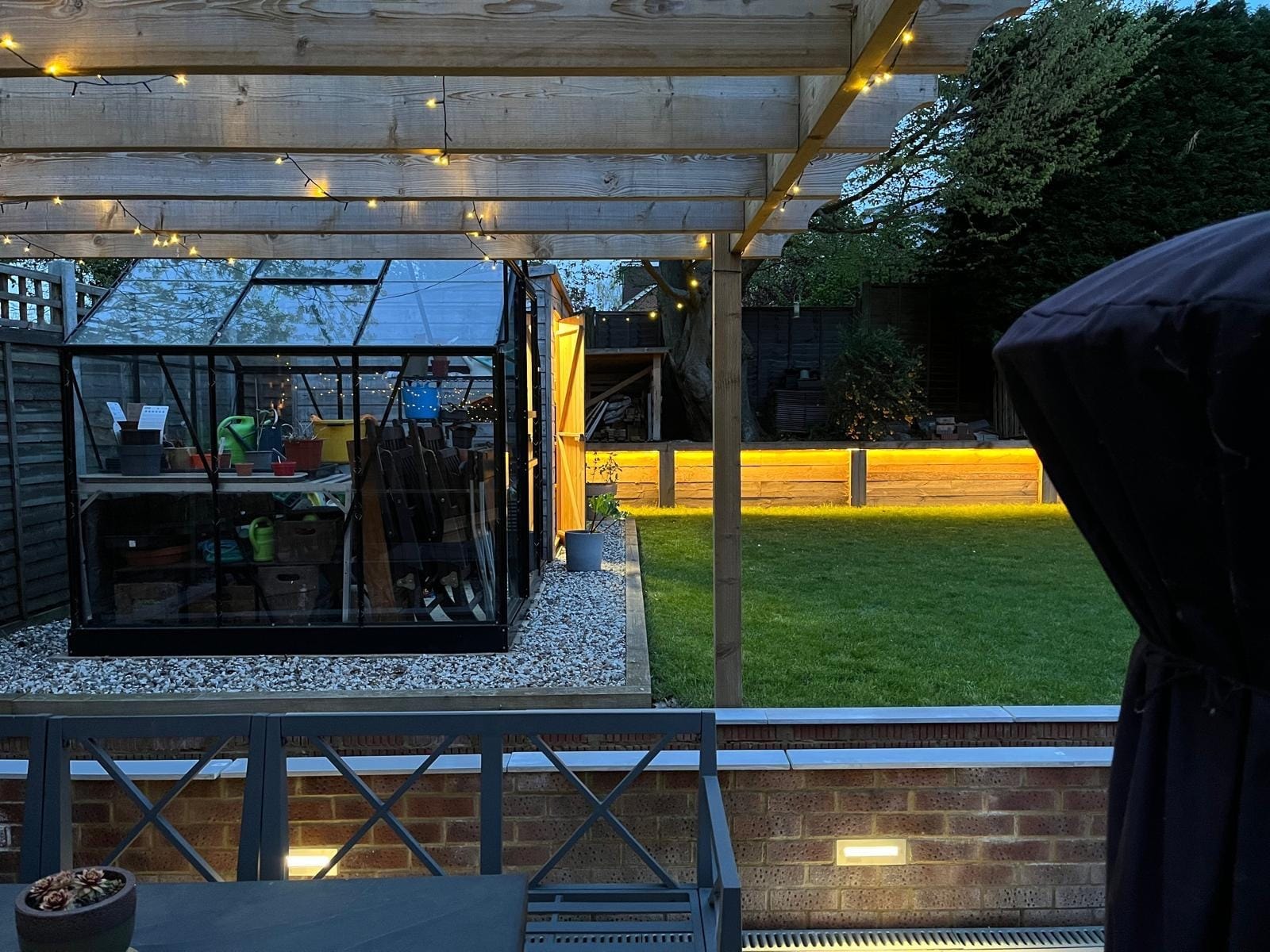 View from the patio under the pergola to the greenhouse, lawn and lit sleeper wall by DNR Build