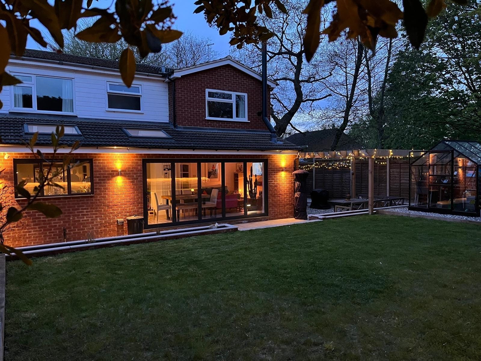 The rear of the house framed by leaves at dusk, with the extension lit and the pergola and greenhouse beside it by DNR Build