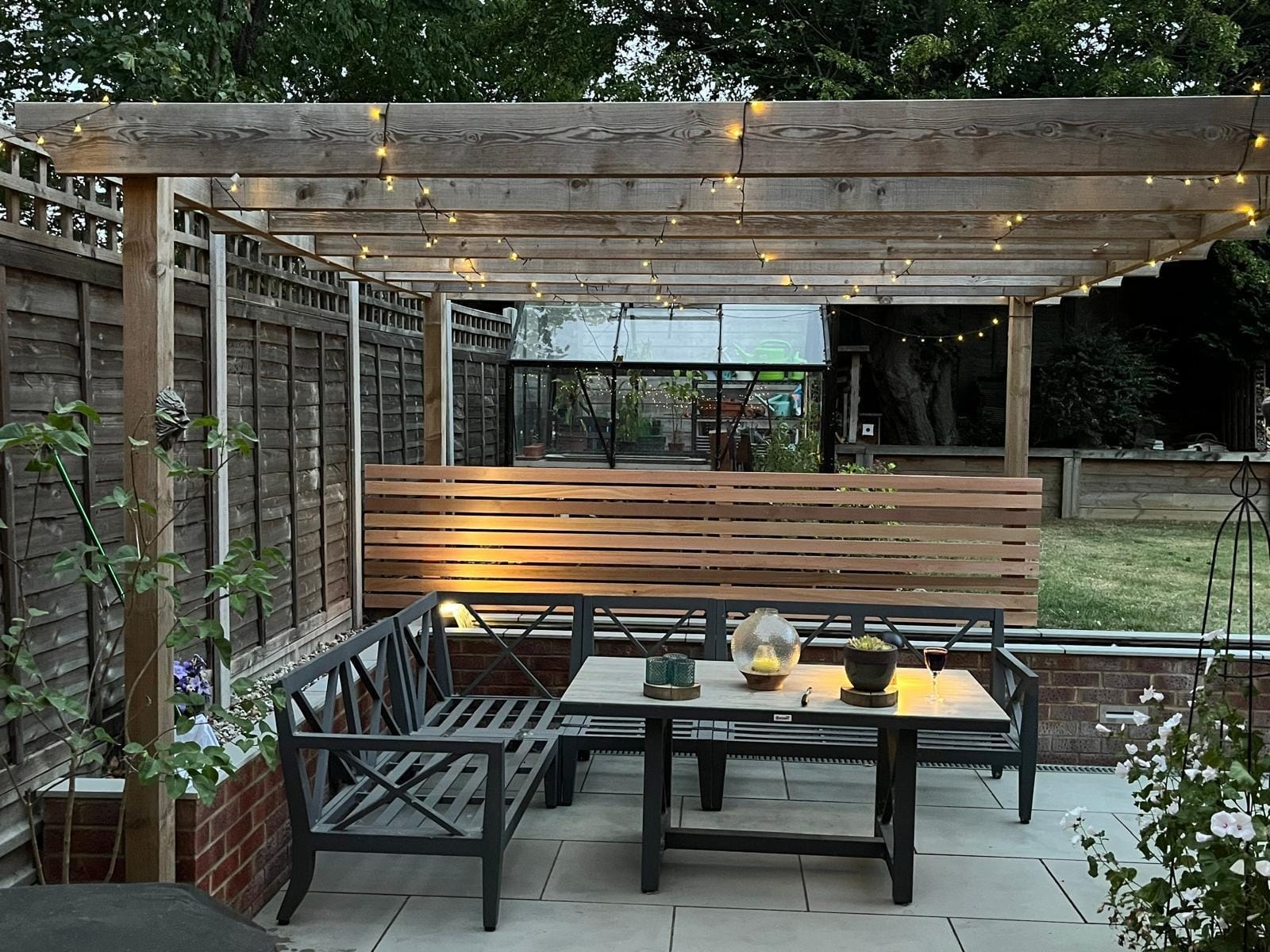 The pergola seating area with festoon lights overhead and a slatted cedar screen behind by DNR Build