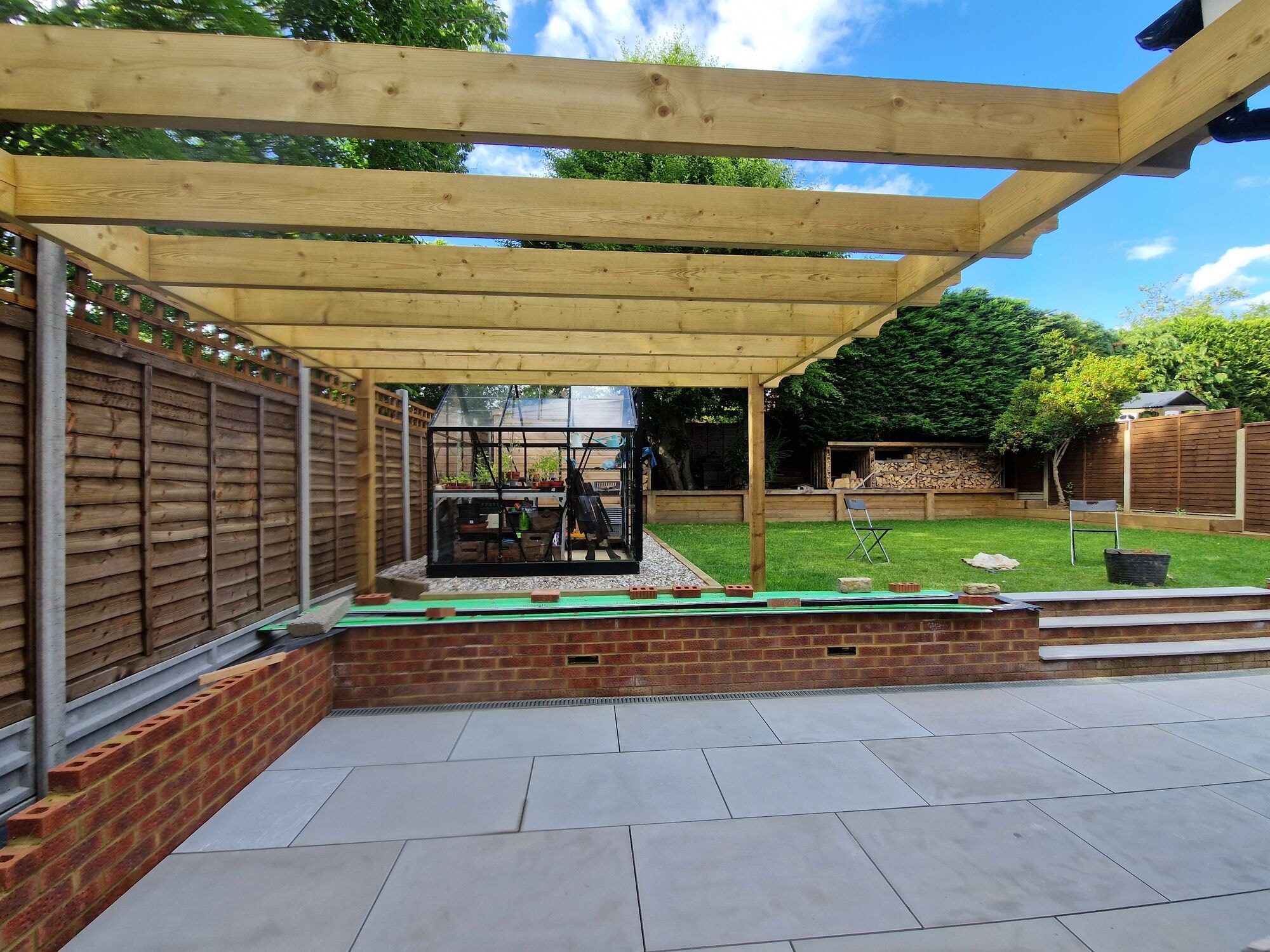 Timber pergola over the patio looking across the lawn to the log stores and greenhouse by DNR Build