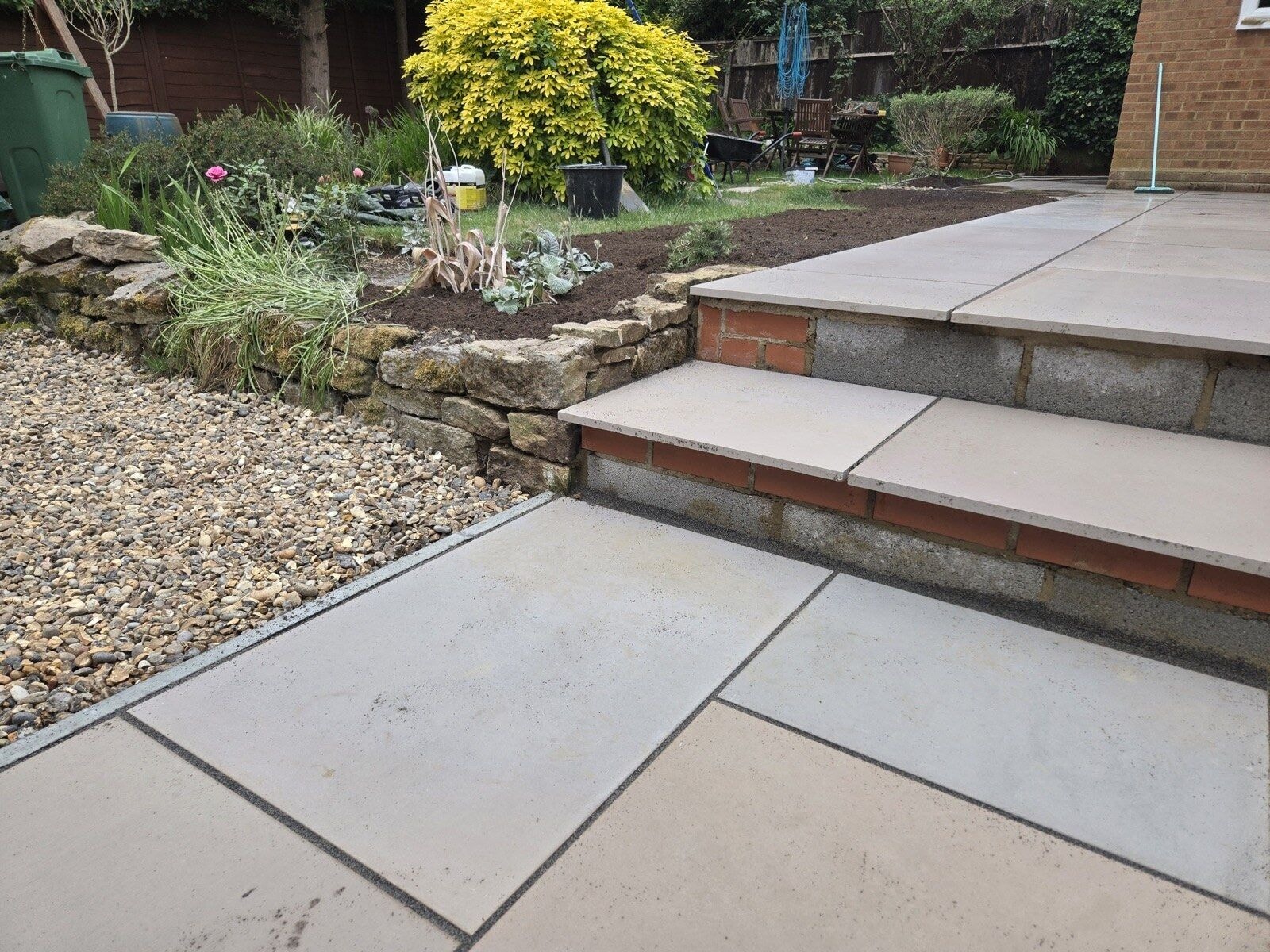 Stepped sandstone patio with brick faced risers leading up from a gravel border and planted bed by DNR Build