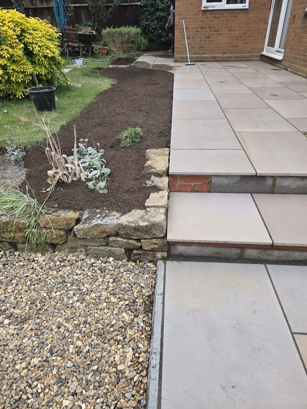 Sandstone patio edge with a reclaimed stone retaining wall and a freshly dug planting bed by DNR Build