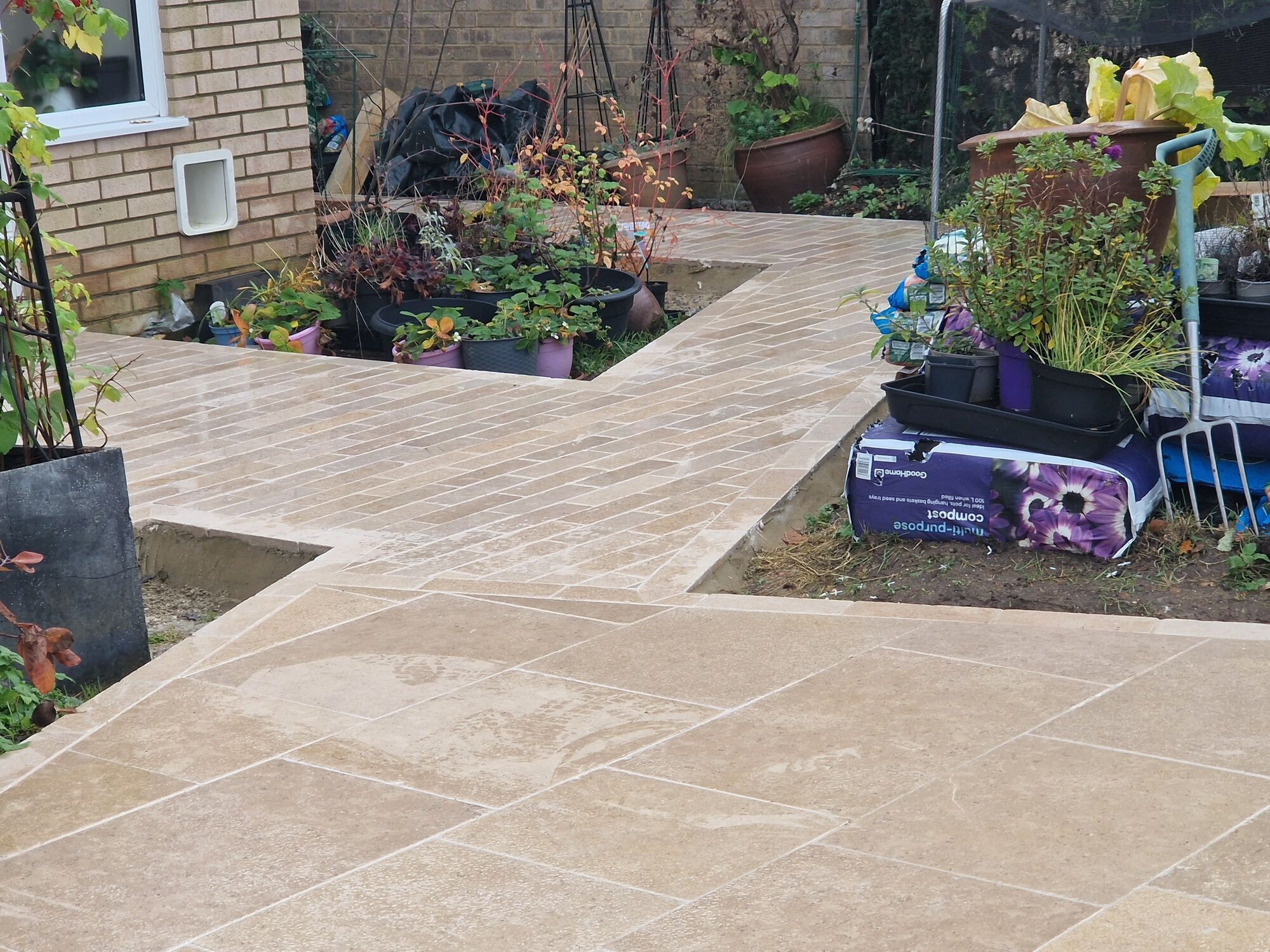 Travertine patio with a plank pattern path leading round past the planted pots by DNR Build